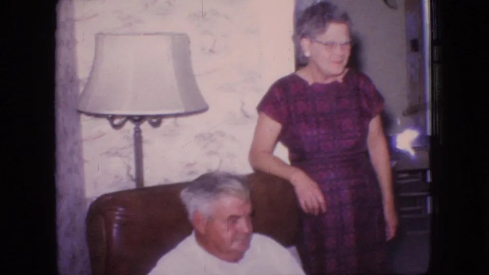 In a 1966 New Jersey home-movie frame, an older man in a white shirt sits in a brown armchair while an older woman in a purple patterned dress and glasses stands nearby beside a floor lamp and floral wallpaper.