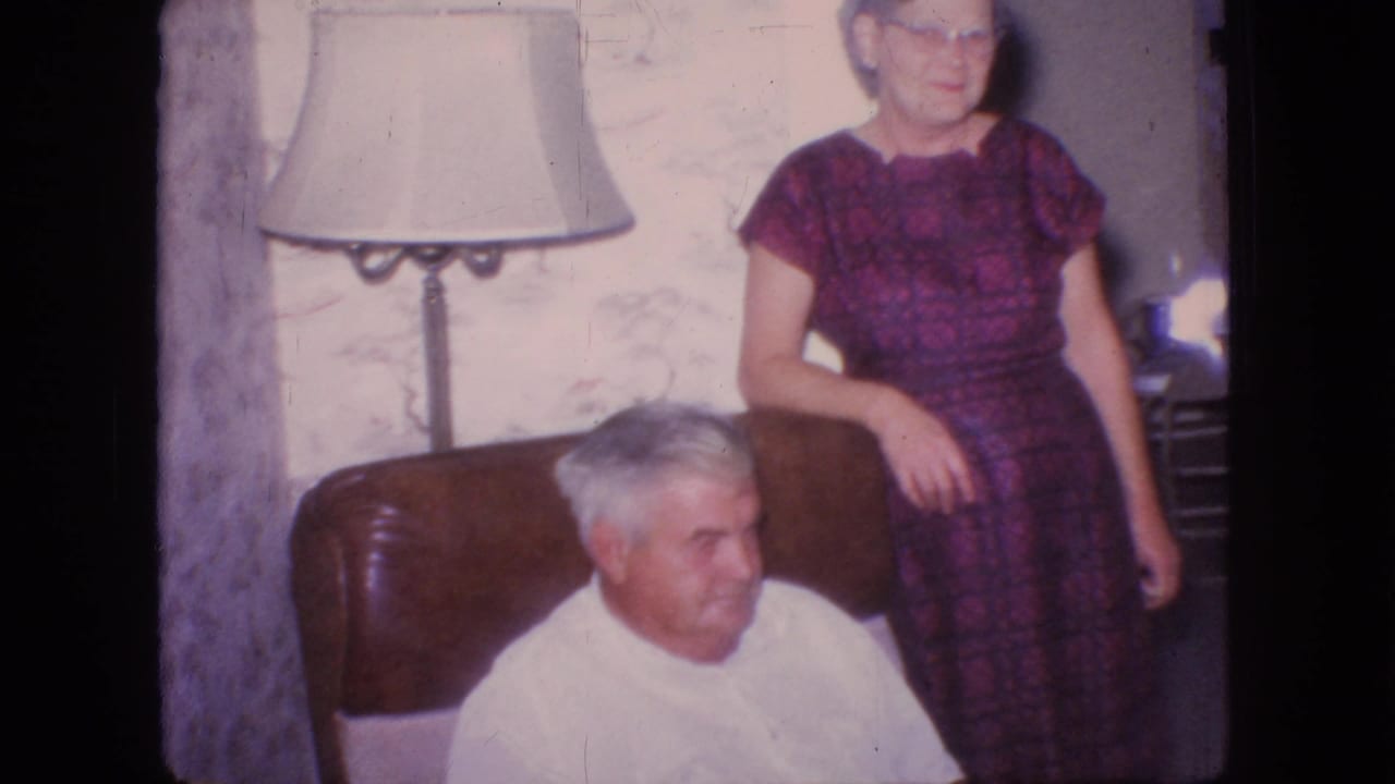 Older woman stands behind a seated older man near a lamp and floral wallpaper.