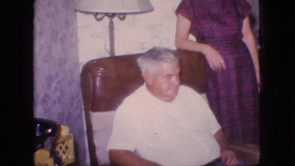 An older man in a white shirt sits in the brown armchair while the standing woman is partly cropped at his side, with the lamp, wallpaper, and home interior details visible.