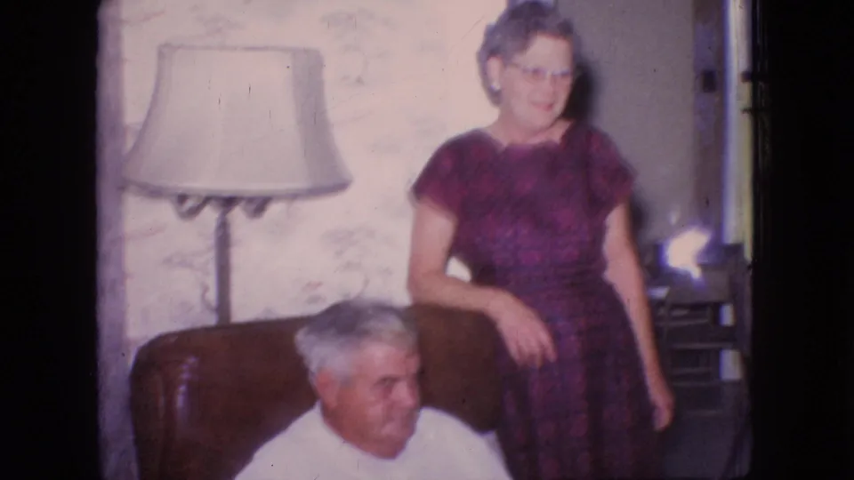 The older woman in a purple patterned dress stands near the chair while the older man sits lower in the foreground, framed by floral wallpaper, a floor lamp, and warm faded 8mm color.