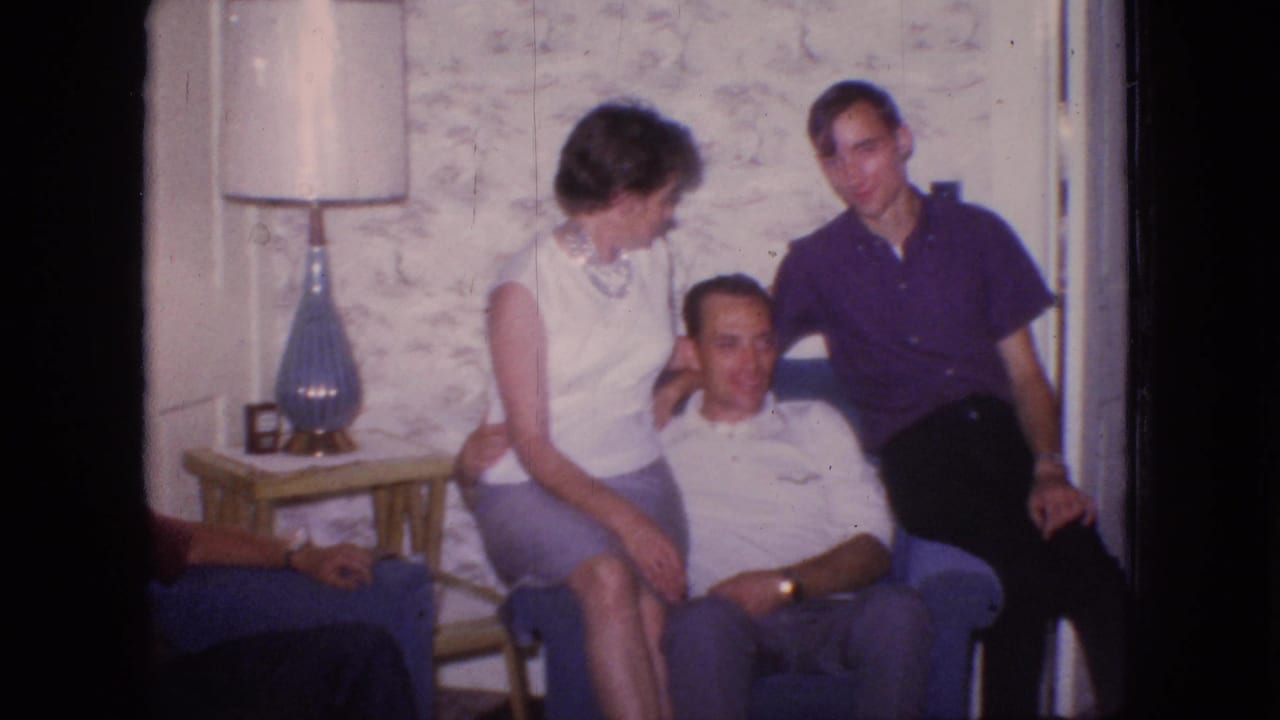 In a 1966 New Jersey home-movie frame, a woman sits on a seated man's lap while a younger man stands beside them in a living room with a blue chair, side table, lamp, and textured wallpaper.