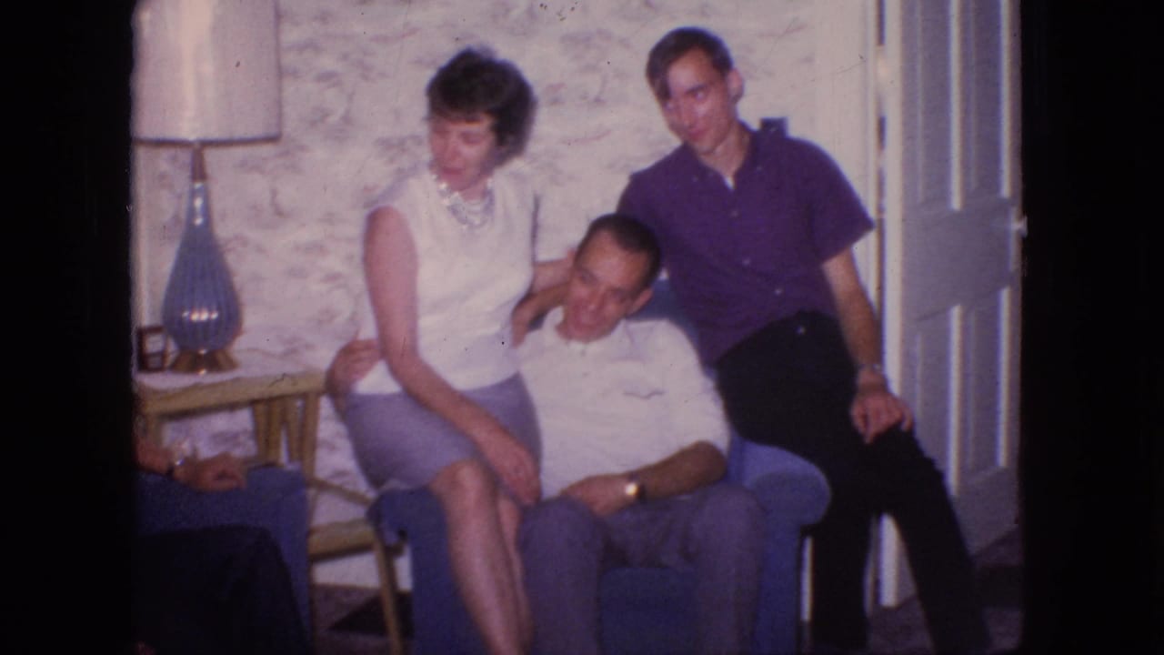 In this 1966 New Jersey living-room frame, a woman in a sleeveless top sits close to a seated man while another man leans in beside an open door, with a lamp and wallpaper behind them.