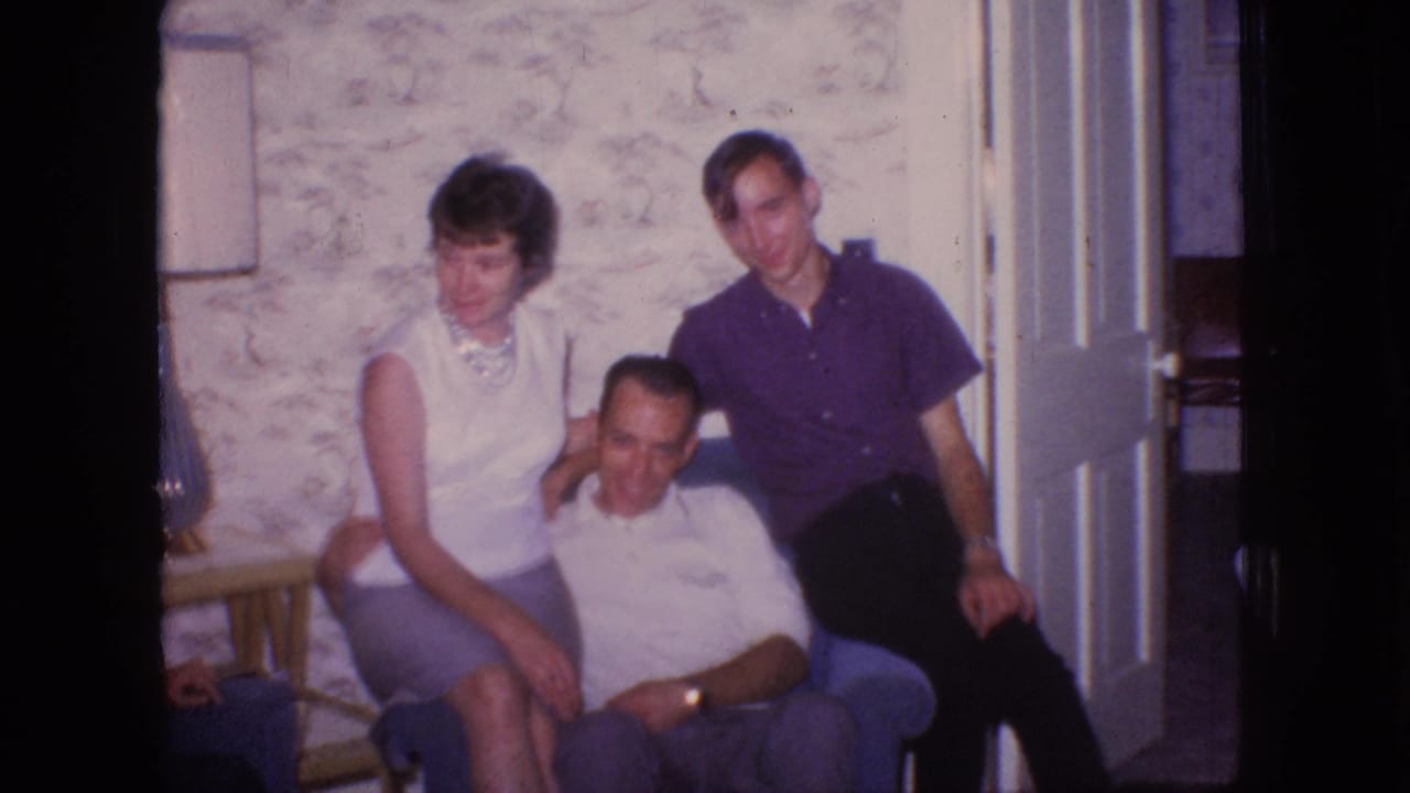 The 1966 New Jersey home-movie frame shows three adults grouped around a blue armchair, with the woman facing the camera, the seated man leaning forward, and a lamp partly visible at the edge.