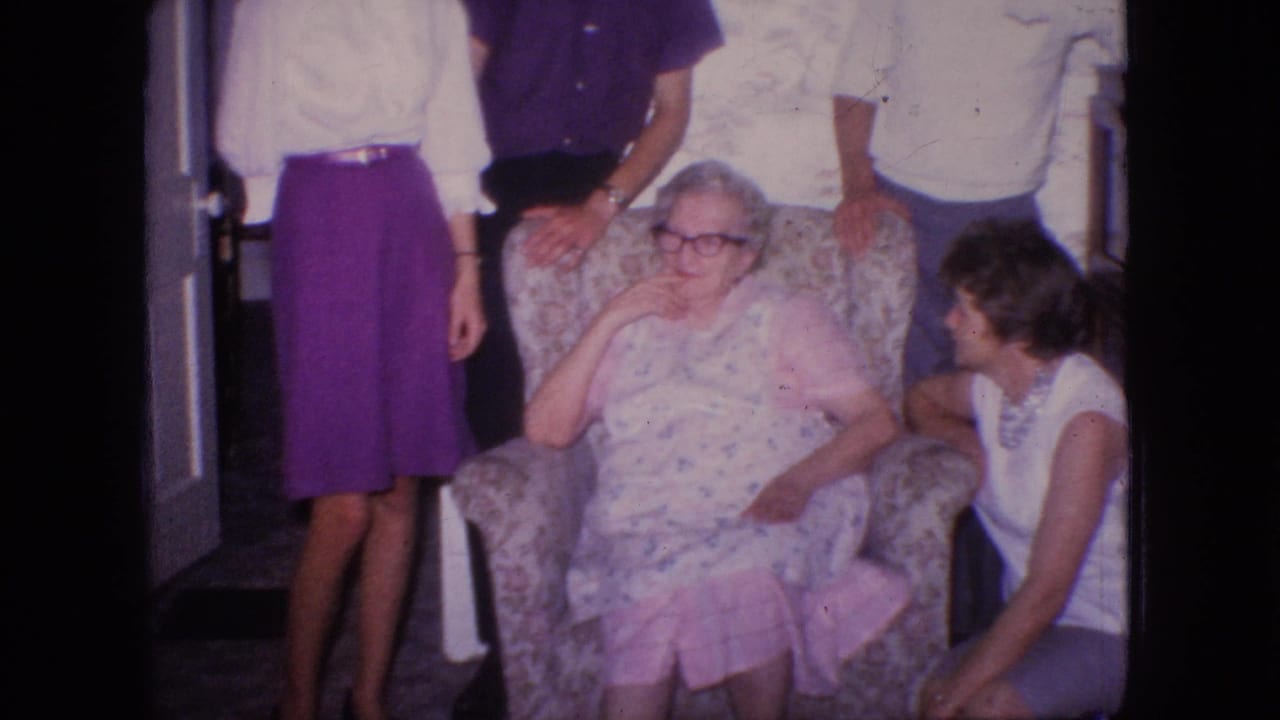 1966 New Jersey home-movie frame showing an elderly woman in glasses and a floral dress seated in a patterned armchair while relatives stand and sit nearby inside a living room.