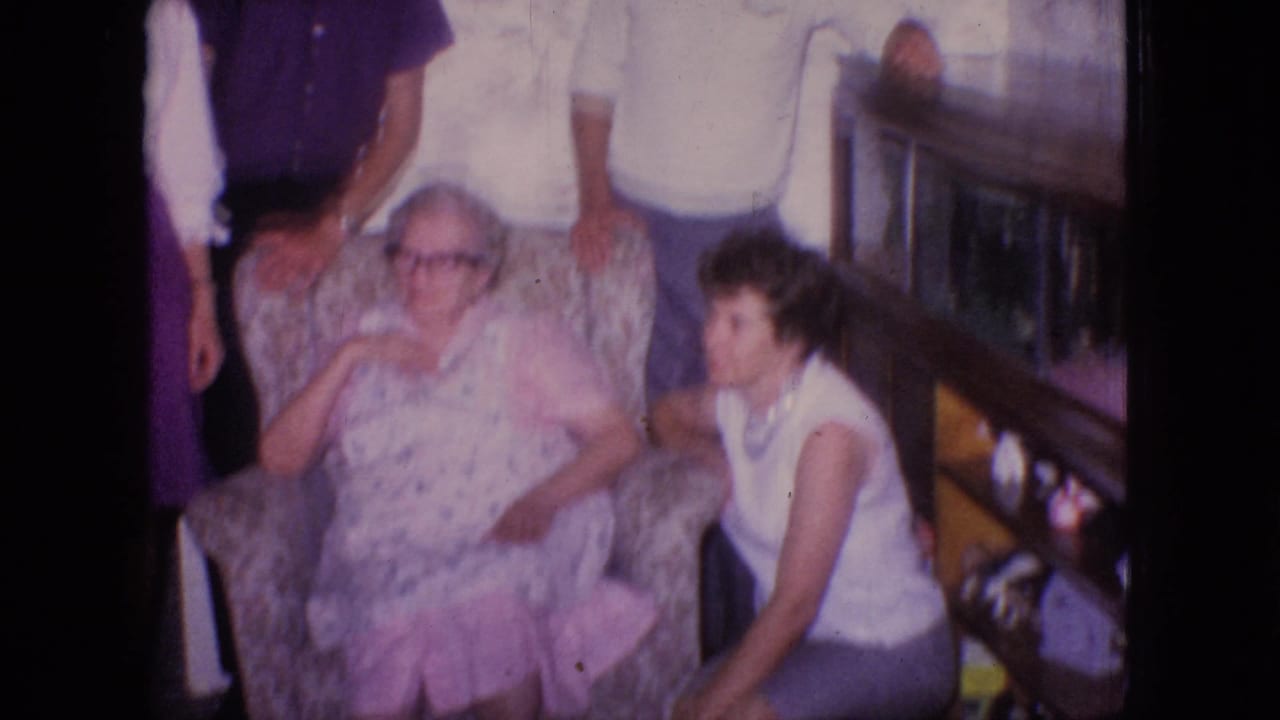 Elderly woman in an armchair with a younger woman seated beside her and relatives standing behind.