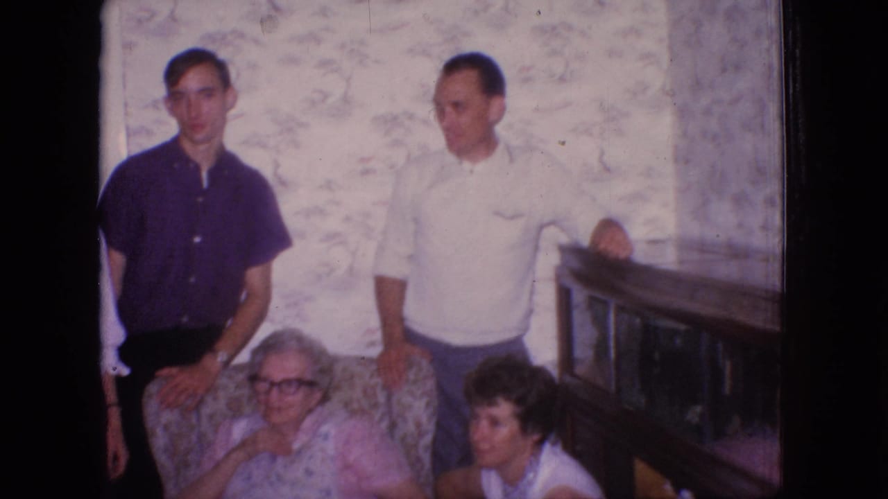 1966 New Jersey home-movie frame showing two men in casual shirts standing behind an elderly woman in a patterned chair, with a younger woman seated near the armrest in a private living room.