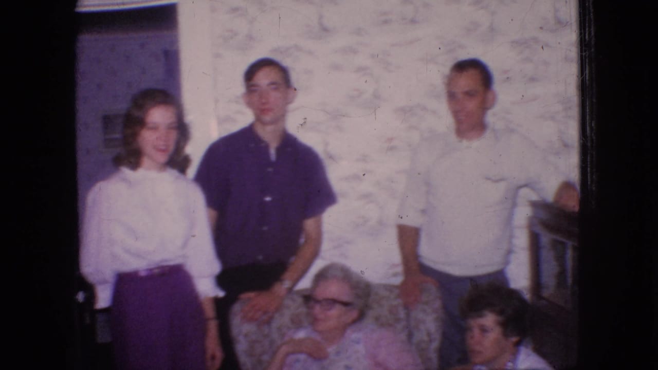 1966 New Jersey home-movie frame of a family group in a living room, including a woman in a white blouse and purple skirt, two standing men, an elderly woman seated in glasses, and a younger seated woman nearby.