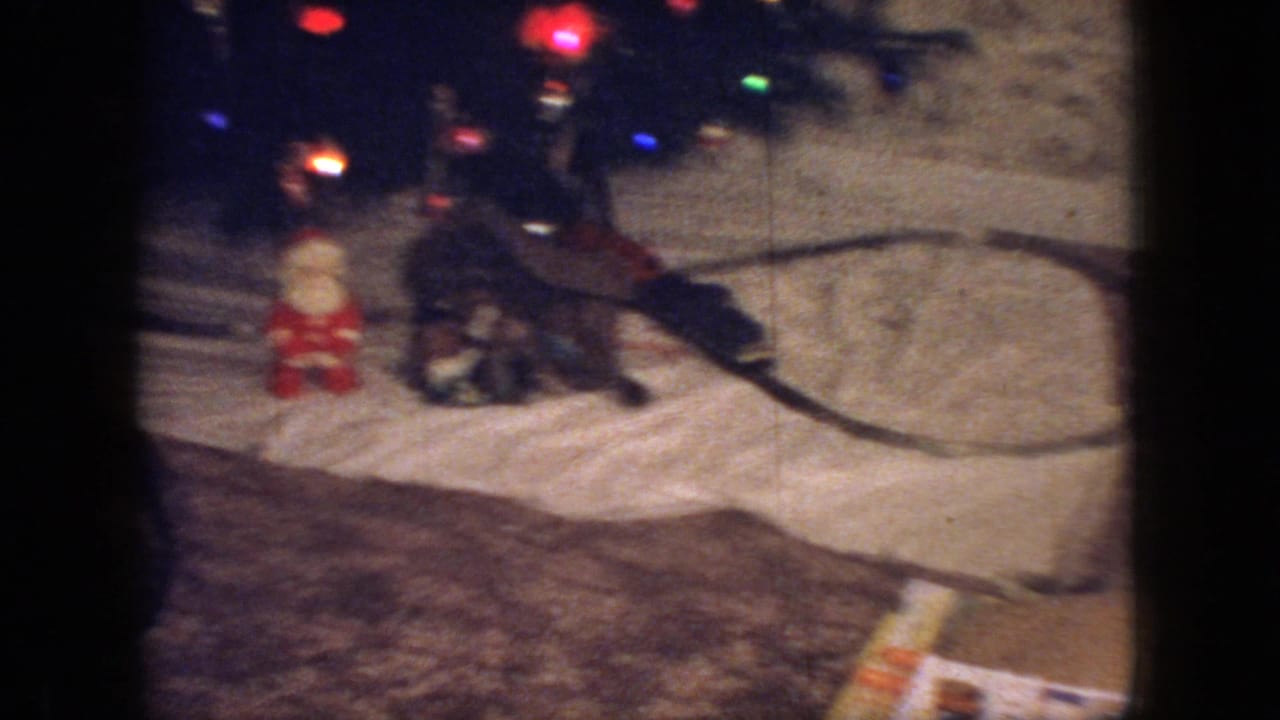 1966 New Jersey home-movie frame showing colored Christmas tree lights, a small Santa figure, and a toy train display arranged on a white blanket inside a private living room.