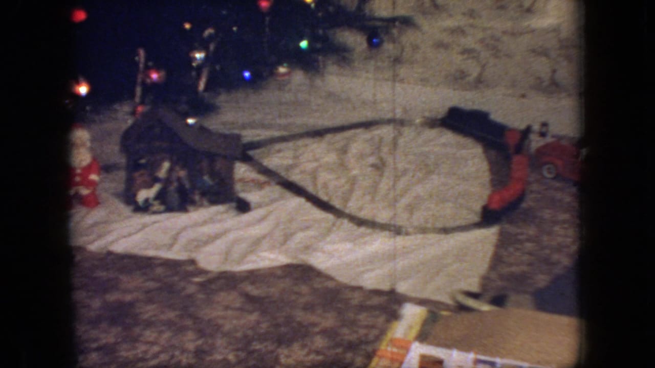 1966 New Jersey home interior frame showing a toy train track loop, small Santa figure, miniature display pieces, and Christmas tree lights in warm 8mm color.