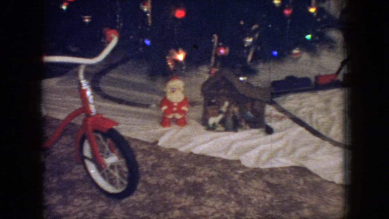1966 New Jersey living-room frame with a red child's tricycle positioned near a lit Christmas tree, a Santa figure, and a toy train display arranged on a blanket.