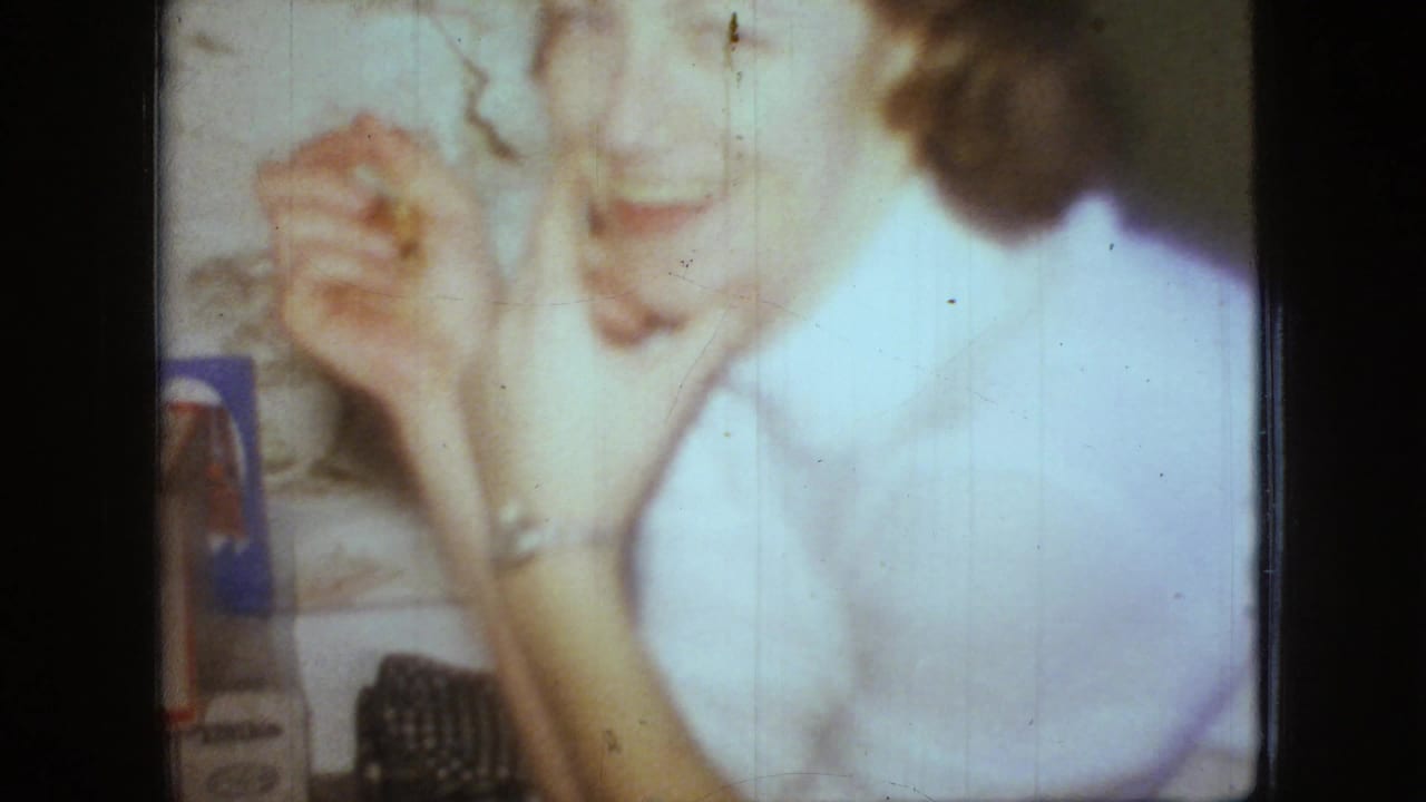 In this 1966 New Jersey home-movie frame, a woman in a light blouse raises her hands near a table while another seated person is partly visible in soft focus.