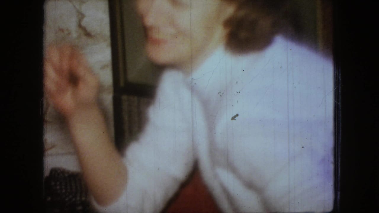 A woman in a light blouse sits at a table in a private interior, her hand lifted near her face as the 8mm frame catches motion and soft indoor light.