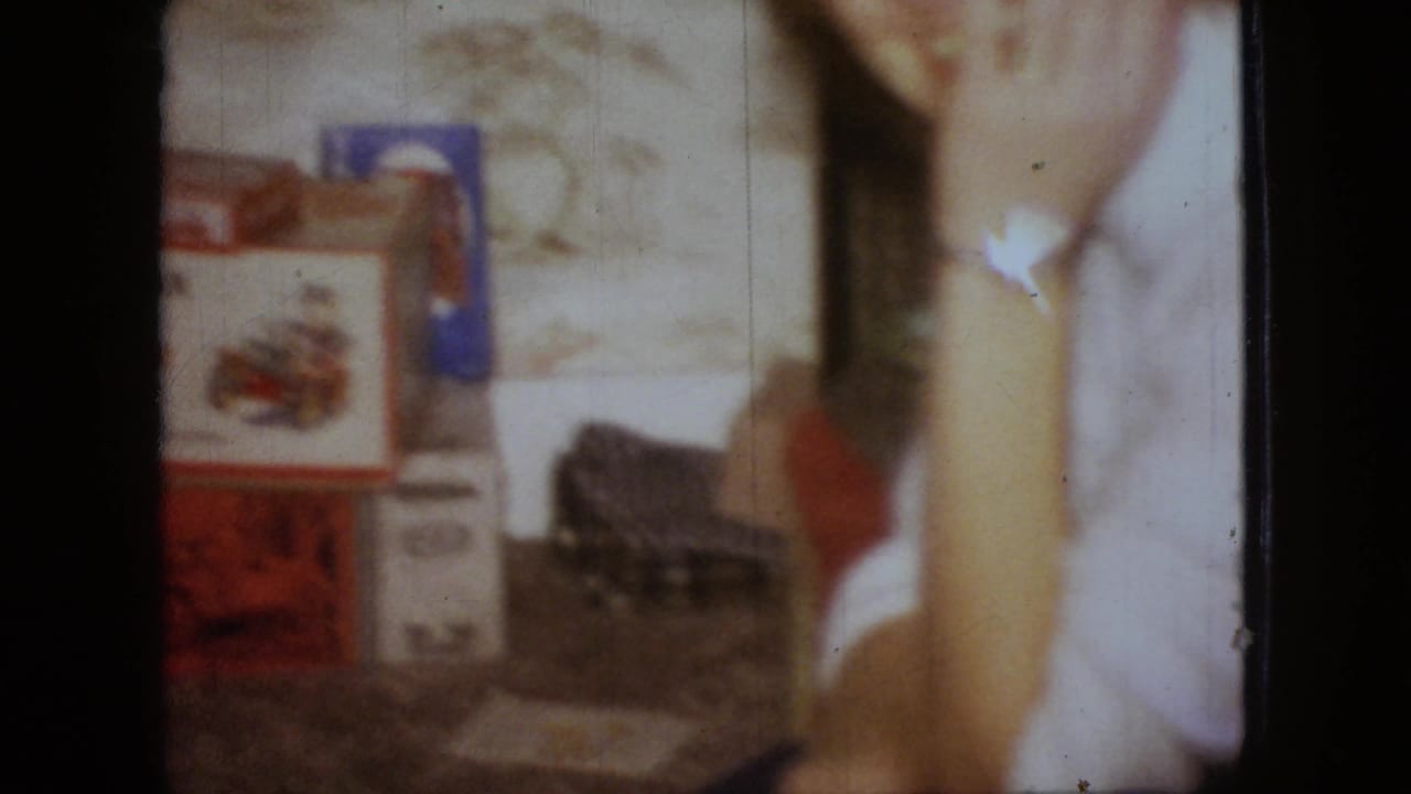 The frame centers on tabletop items and a woman's arm, with boxed or packaged objects visible against a floral interior background in the 1966 home-movie scene.