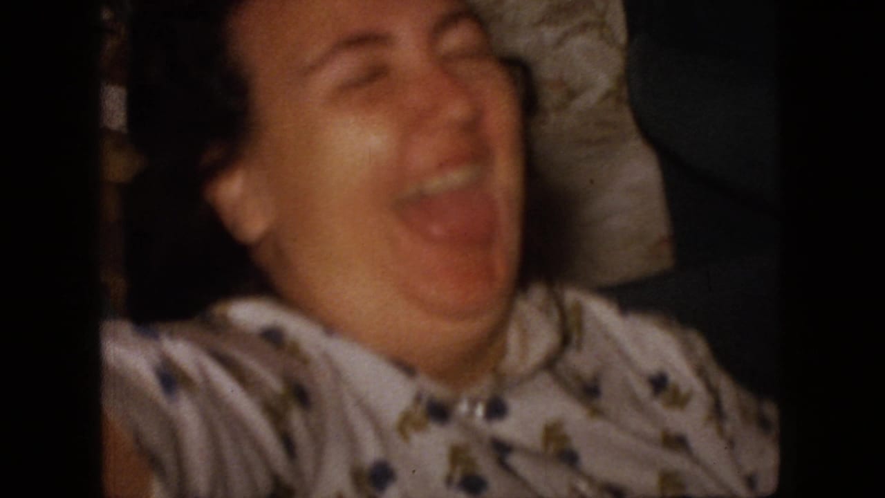 A woman in a patterned blouse laughs broadly in a close, softly focused 1966 New Jersey home-movie frame, with a dark partial figure at the edge of the private interior.