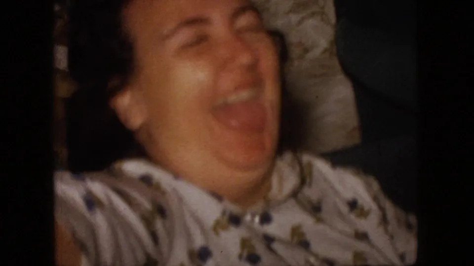 A woman in a patterned blouse laughs broadly in a close, softly focused 1966 New Jersey home-movie frame, with a dark partial figure at the edge of the private interior.