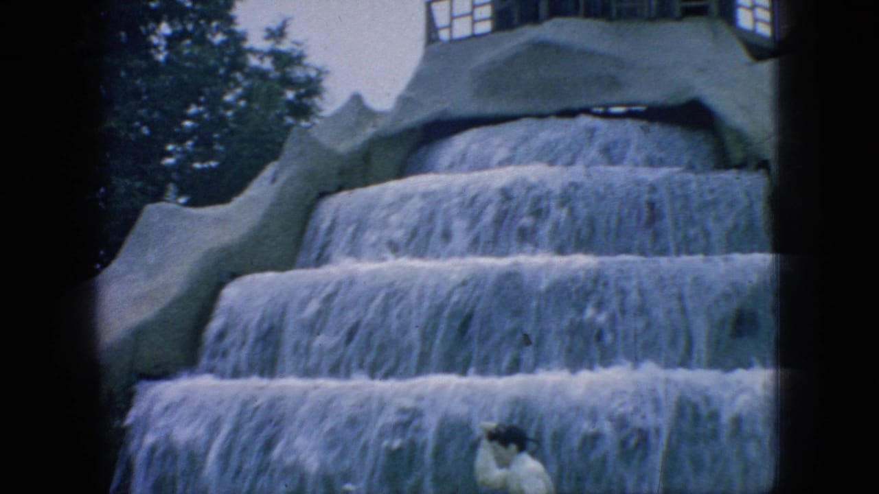 1966 New Jersey 8mm frame showing a man partly visible on grass before a multi-tiered artificial waterfall display, with trees and dark film-frame edges around the scene.