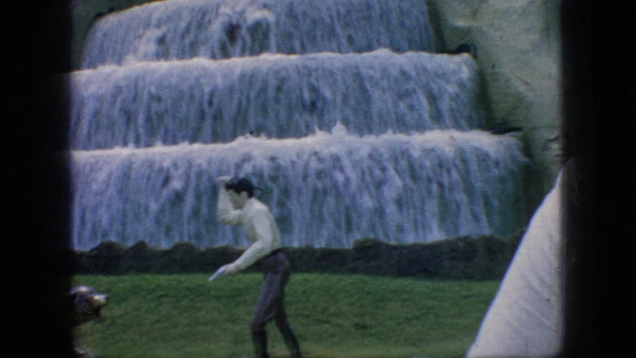 1966 New Jersey 8mm frame showing a man walking across a grassy area near an animal display, with artificial waterfalls filling the background.