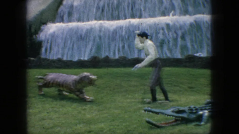 1966 New Jersey home-movie frame showing a man gesturing near a striped tiger figure and large alligator form on grass in front of a tiered artificial waterfall.