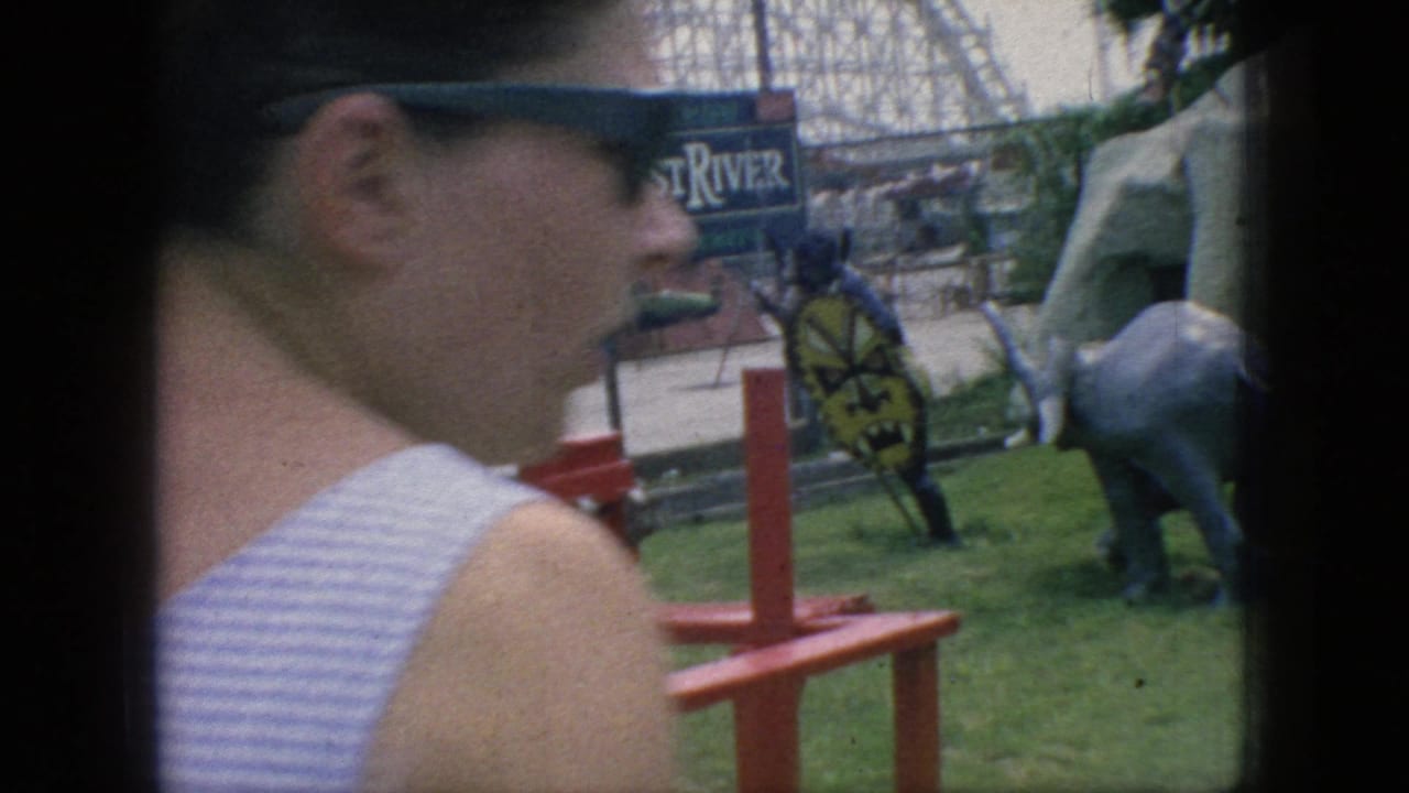 A park visitor in sunglasses and sleeveless striped clothing passes in the foreground as the Lost River sign, red ride structure, elephant statue, and artificial rockwork remain visible behind her.