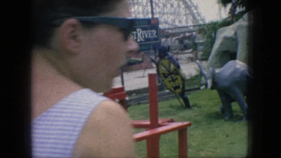 A park visitor in sunglasses and sleeveless striped clothing passes in the foreground as the Lost River sign, red ride structure, elephant statue, and artificial rockwork remain visible behind her.