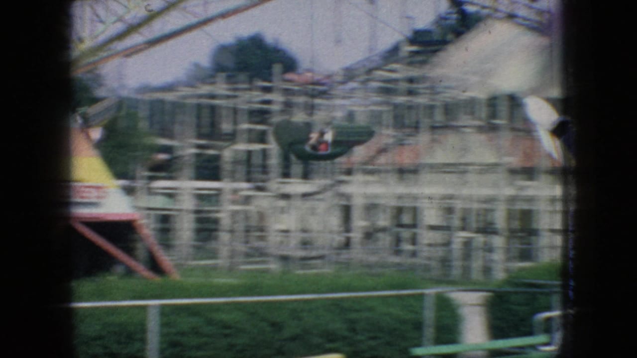 1966 New Jersey 8mm frame showing a ride car moving past a pale building, dense metal railings, lawn edging, and partial amusement-ride structures.