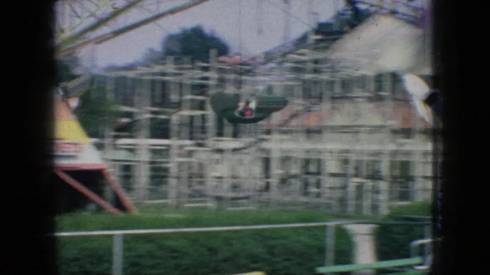 1966 New Jersey 8mm frame showing a ride car moving past a pale building, dense metal railings, lawn edging, and partial amusement-ride structures.