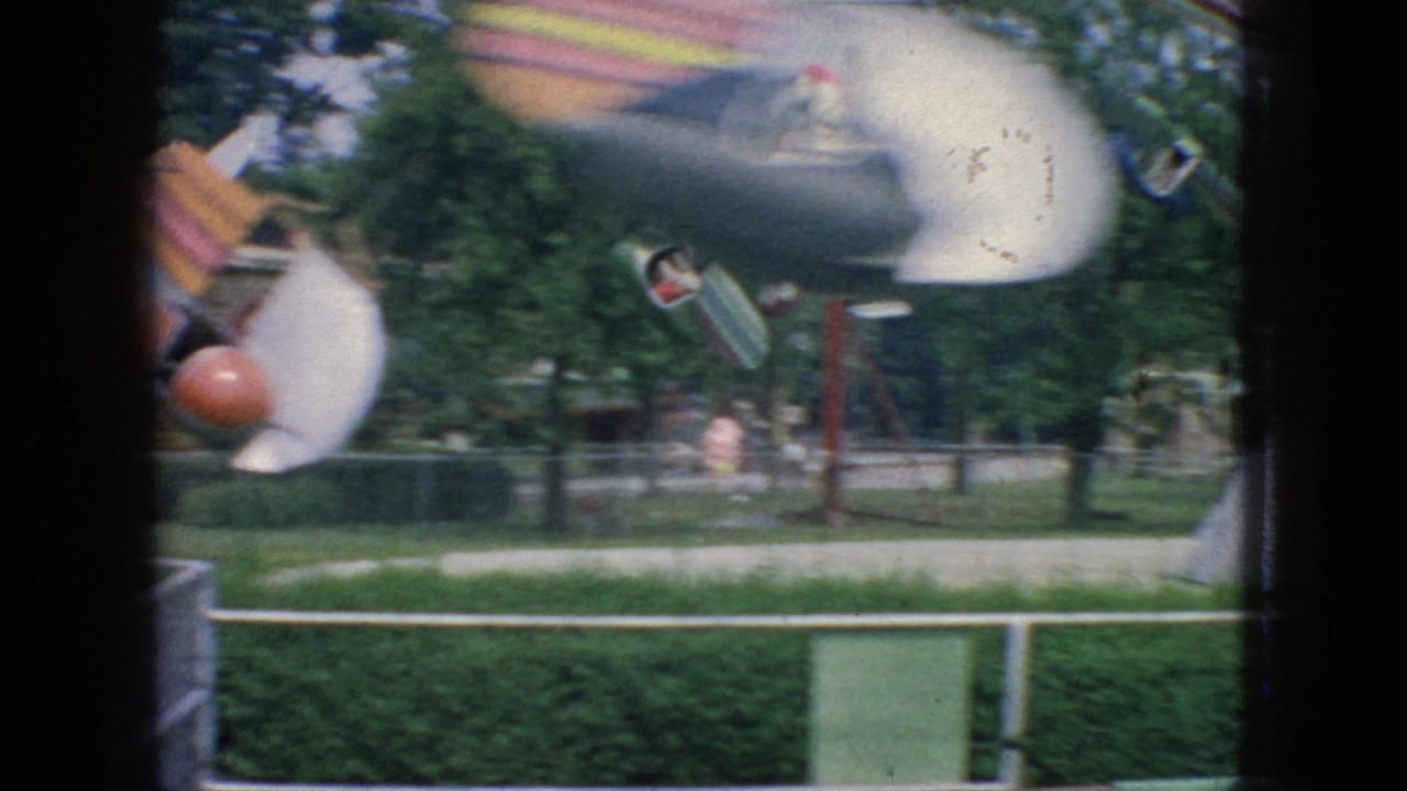 1966 New Jersey 8mm frame showing an airplane-style rotating attraction with suspended cars, visible riders, grass, railings, and trees in the background.