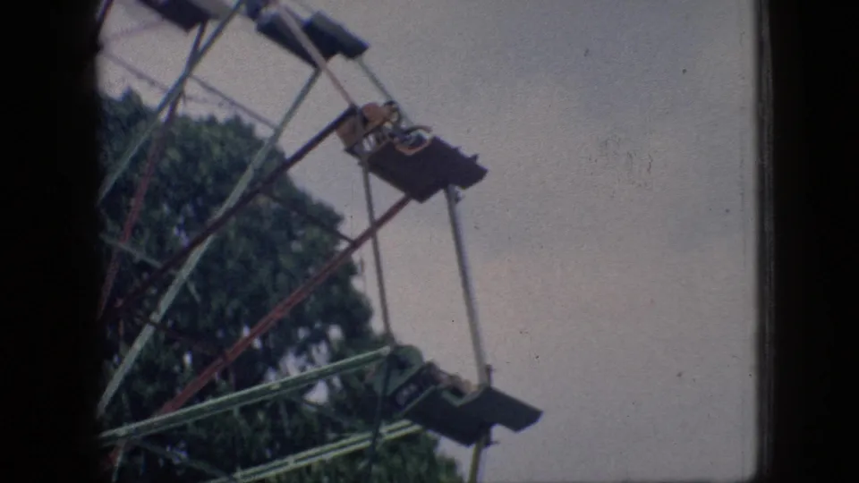 1966 New Jersey home-movie frame showing part of a Ferris wheel with suspended seats, riders at a distance, trees, pale sky, and dark film-edge borders.