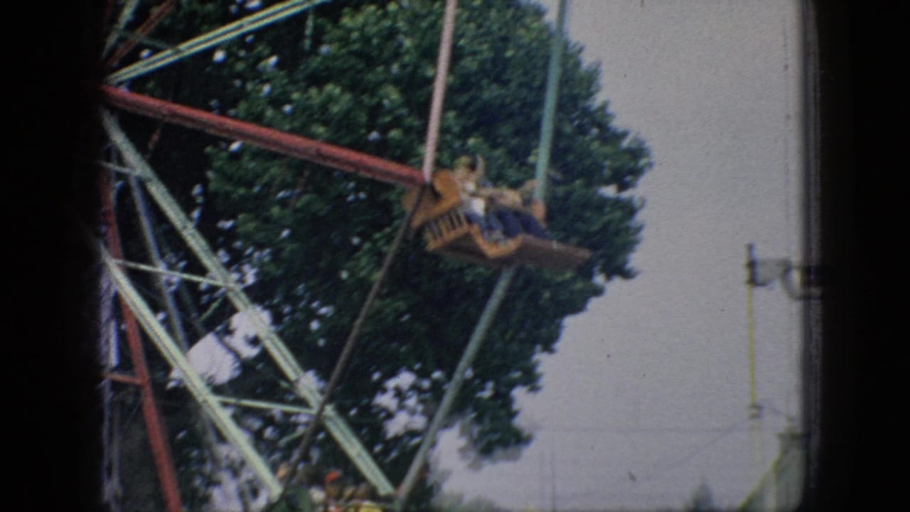 A Ferris wheel gondola carrying distant riders moves across the upper frame, with green trees behind the painted metal ride structure.