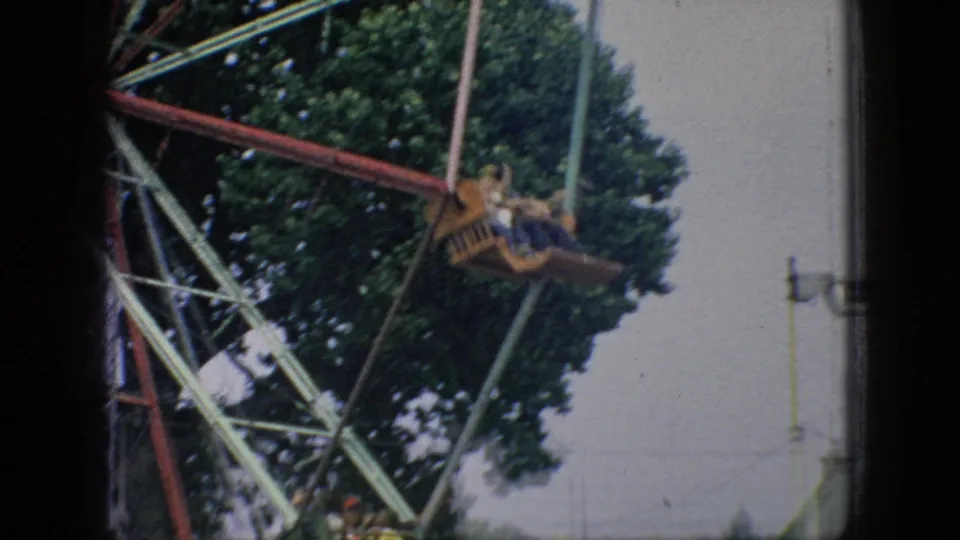 A Ferris wheel gondola carrying distant riders moves across the upper frame, with green trees behind the painted metal ride structure.