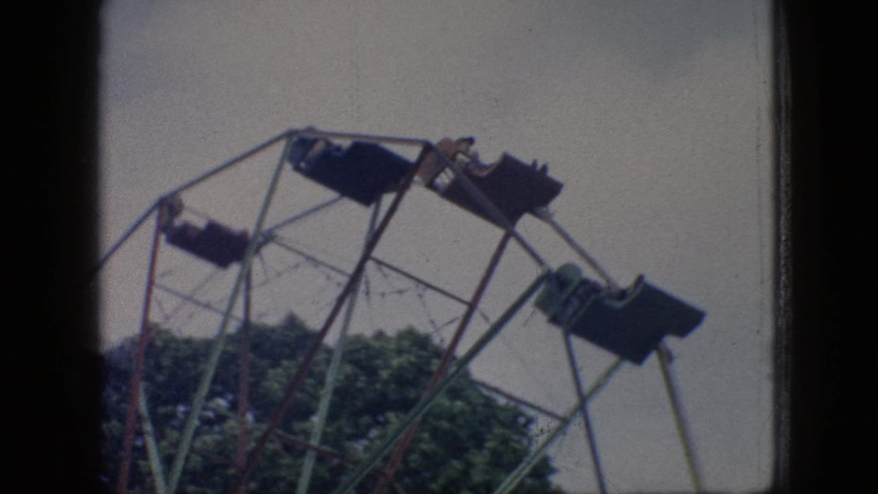 The Ferris wheel structure fills the frame with multiple suspended seats, distant riders, trees below, and the soft color and grain of scanned 8mm film.
