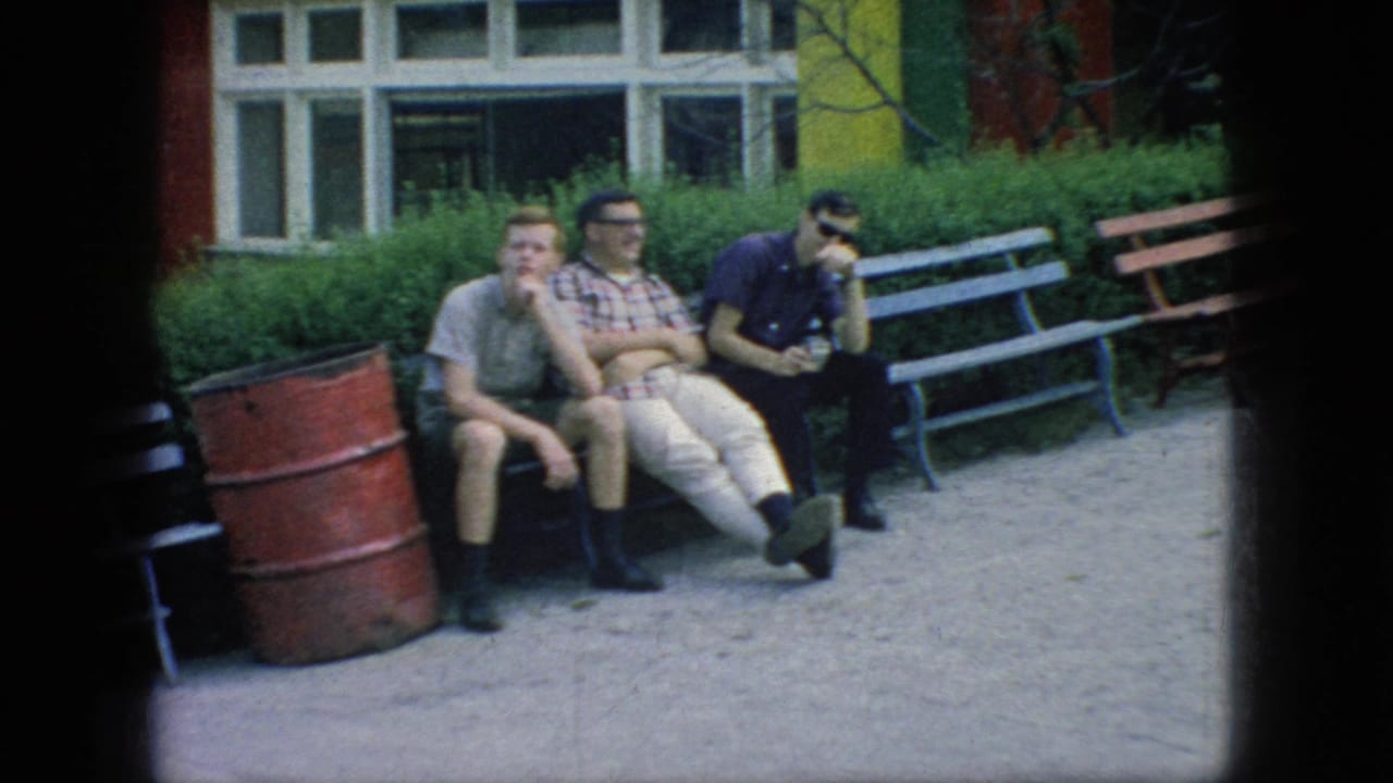 Three young men in casual 1960s clothing sit on a bench at a New Jersey fairground, with a red barrel, painted benches, shrubs, and a building behind them.