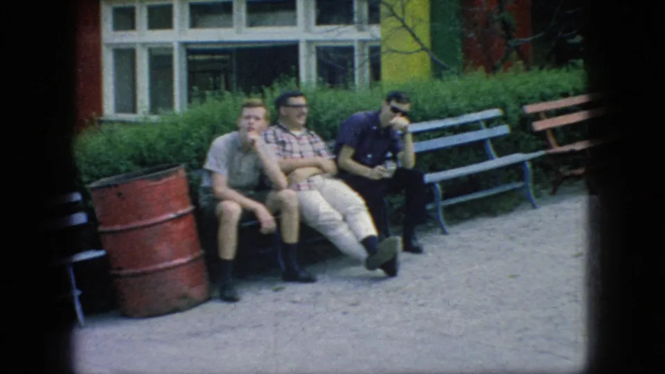Three young men in casual 1960s clothing sit on a bench at a New Jersey fairground, with a red barrel, painted benches, shrubs, and a building behind them.