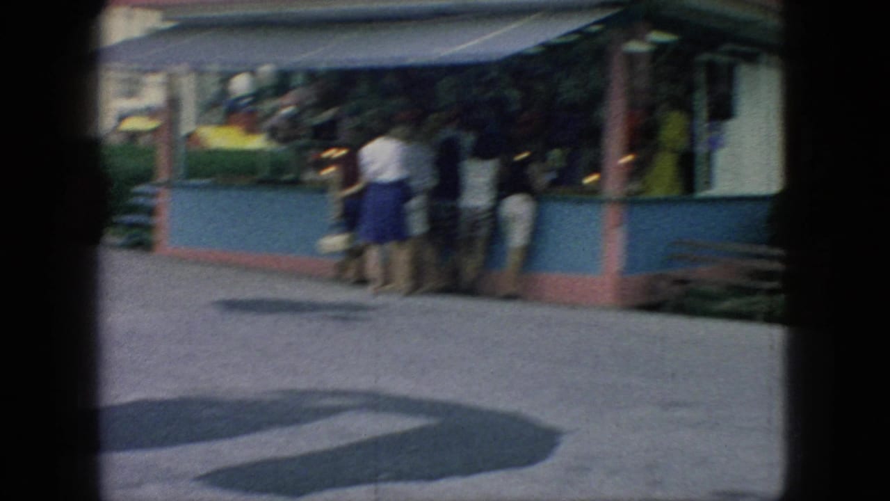 Handheld 8mm footage shows a slightly soft view of fair visitors gathered at a concession stand, with a paved open area in the foreground.