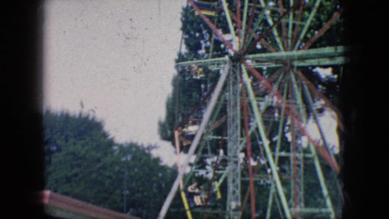 1966 New Jersey 8mm frame showing part of a colorful Ferris wheel with red and green supports, leafy trees, pale sky, and a heavy dark scan border.