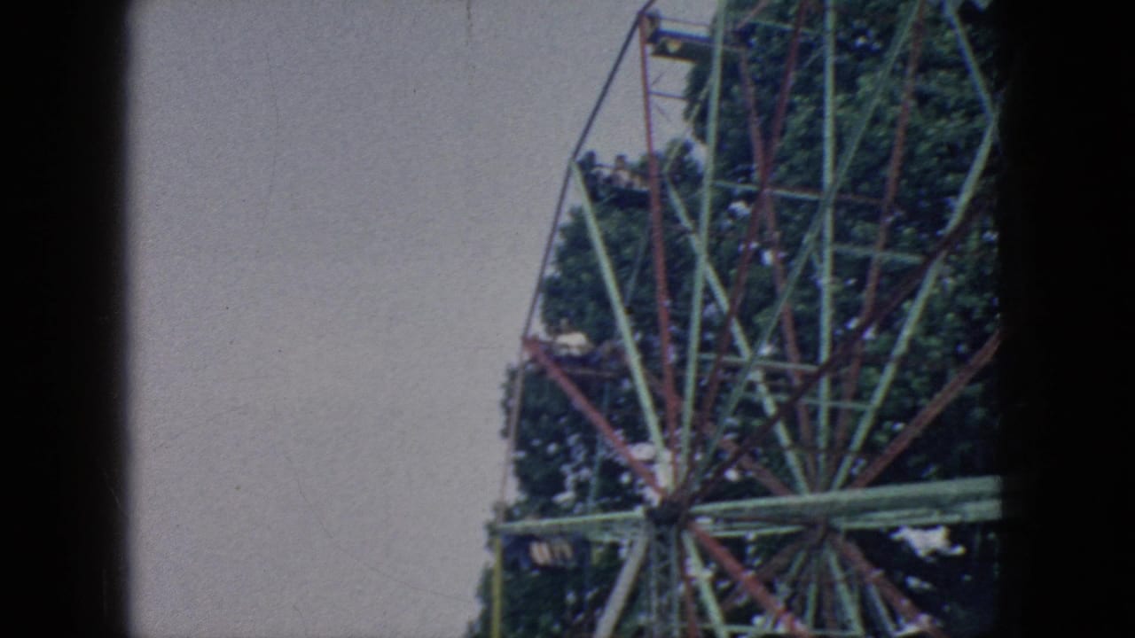 1966 New Jersey 8mm frame showing the upper right portion of a Ferris wheel, with suspended cars, painted metal framework, trees, pale sky, and edge darkening from the scan.