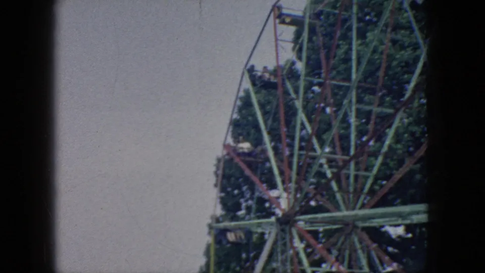 1966 New Jersey 8mm frame showing the upper right portion of a Ferris wheel, with suspended cars, painted metal framework, trees, pale sky, and edge darkening from the scan.
