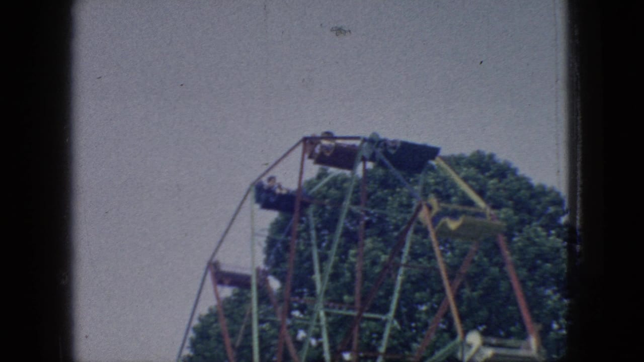1966 New Jersey 8mm frame showing Ferris wheel cars and angular red and green supports rotating in front of leafy trees under a pale sky.