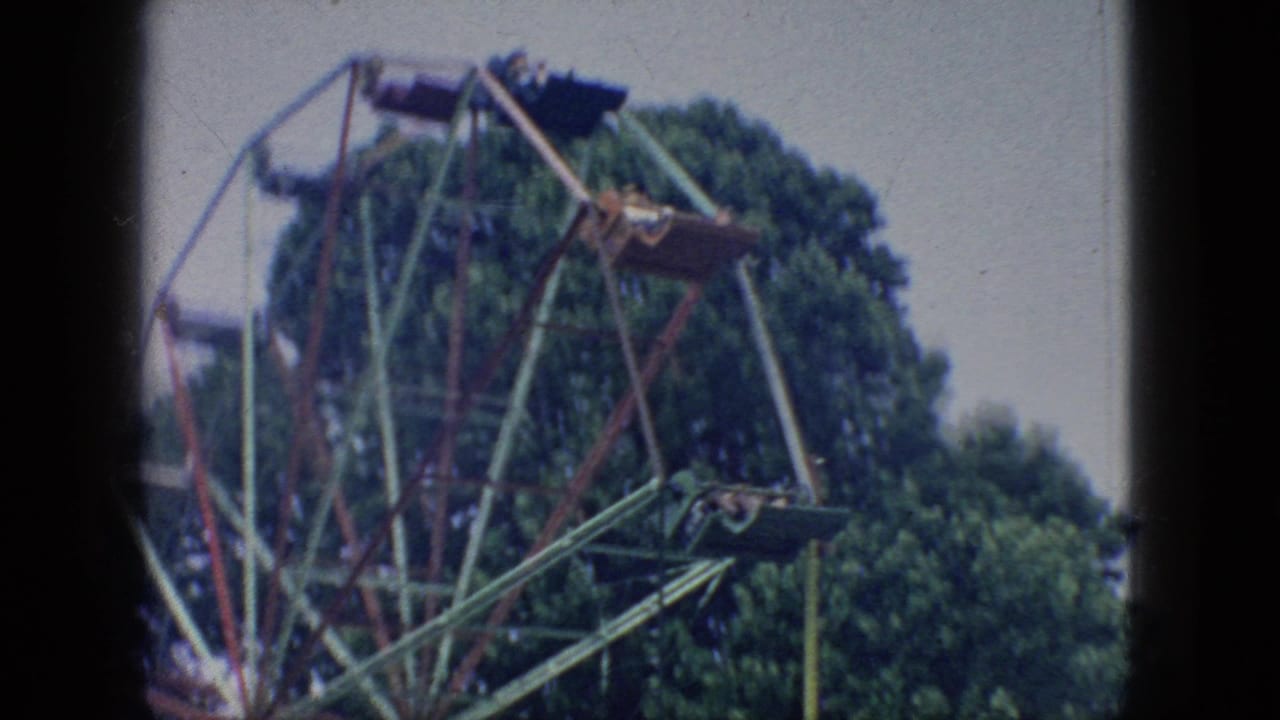 1966 New Jersey 8mm frame showing a closer view of the Ferris wheel structure, several suspended cars with distant riders, leafy trees, and visible film softness.