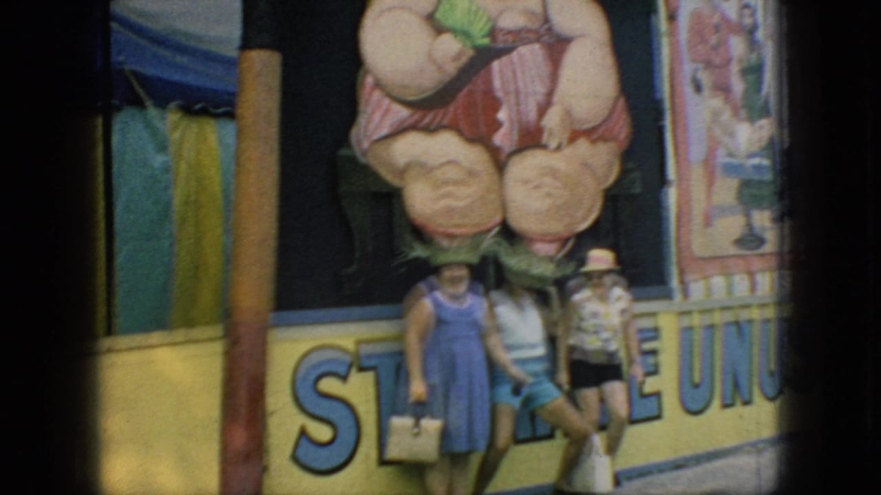 In 1966 New Jersey home-movie footage, three women in summer clothing pose before a carnival booth decorated with a large painted figure and bold sign lettering.