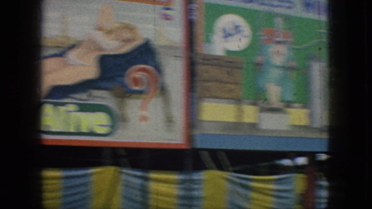 The frame shows carnival booth paintings with stylized figures, partial text, and bright striped trim in a soft, grainy 8mm home-movie view.