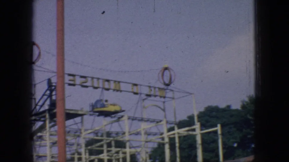 1966 New Jersey amusement park frame showing a small roller coaster track, a car on the elevated structure, overhead wires, trees, and partial ride signage.