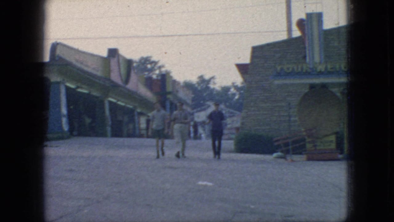 Three young men walk through a 1966 New Jersey amusement park walkway past low attraction or concession buildings, trees, and a masonry facade in soft 8mm home-movie footage.