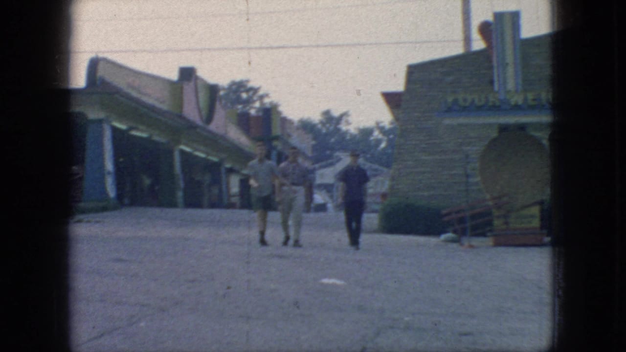 Three young men continue along the paved walkway at a 1966 New Jersey amusement park, passing a low building with partial signage and open pedestrian space.