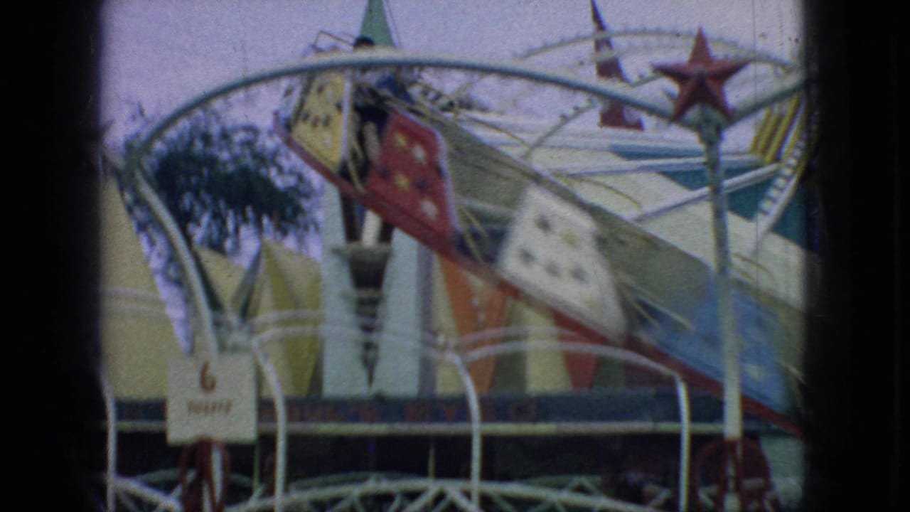 1966 New Jersey 8mm frame showing the carnival ride from a closer angle, with star-painted cars, white railings, a small ticket sign, and a palm-like decoration nearby.