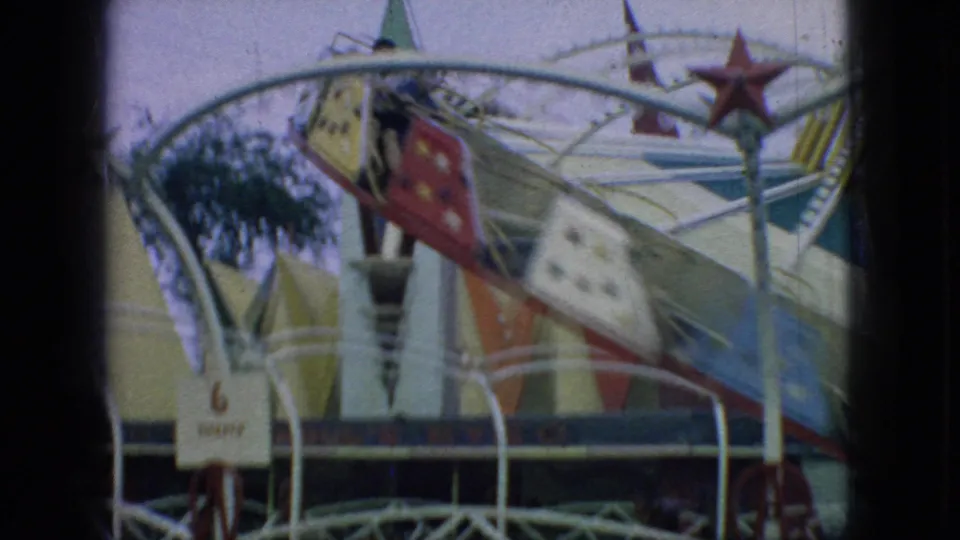 1966 New Jersey 8mm frame showing the carnival ride from a closer angle, with star-painted cars, white railings, a small ticket sign, and a palm-like decoration nearby.