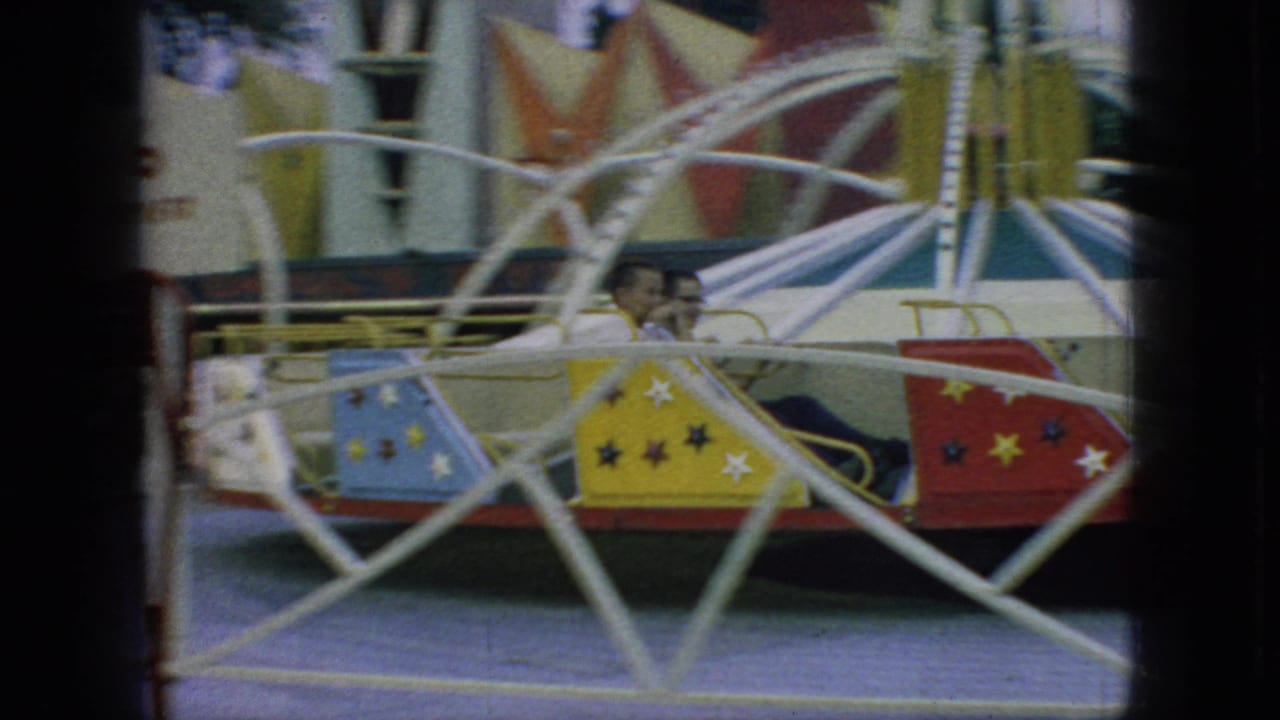 1966 New Jersey home-movie frame with visible riders seated in blue, yellow, and red star-decorated carnival swing cars behind white curved safety rails.