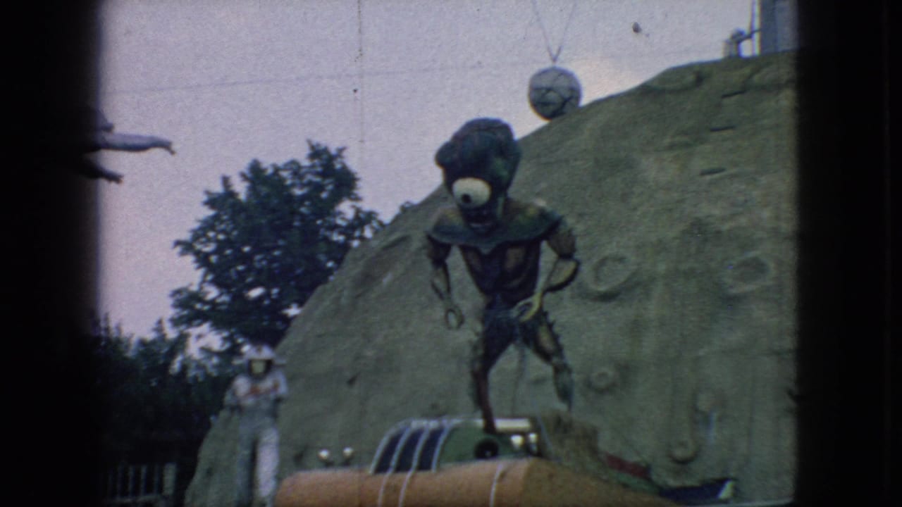 1966 New Jersey 8mm frame showing a one-eyed alien figure over an orange rocket-like attraction, with a white astronaut figure beside a cratered backdrop.