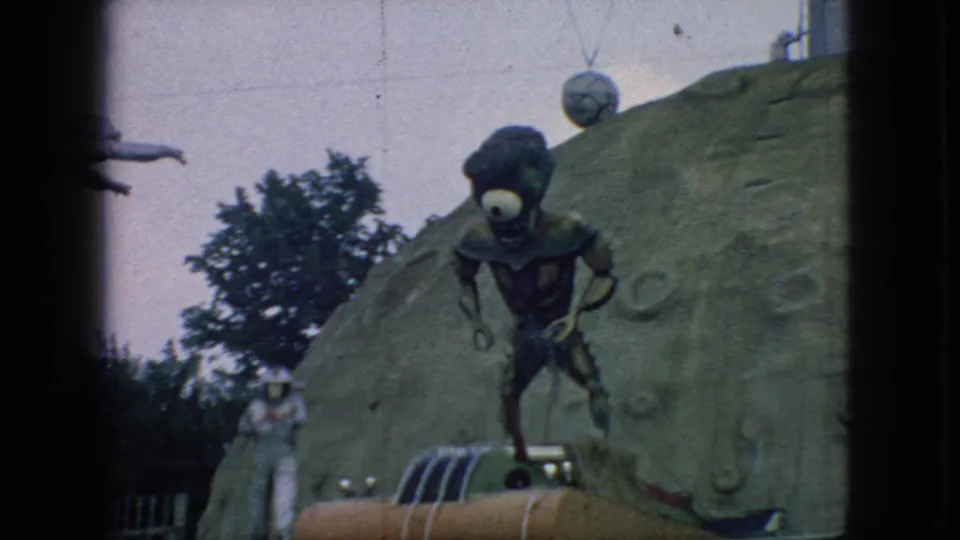 1966 New Jersey 8mm frame showing a one-eyed alien figure over an orange rocket-like attraction, with a white astronaut figure beside a cratered backdrop.