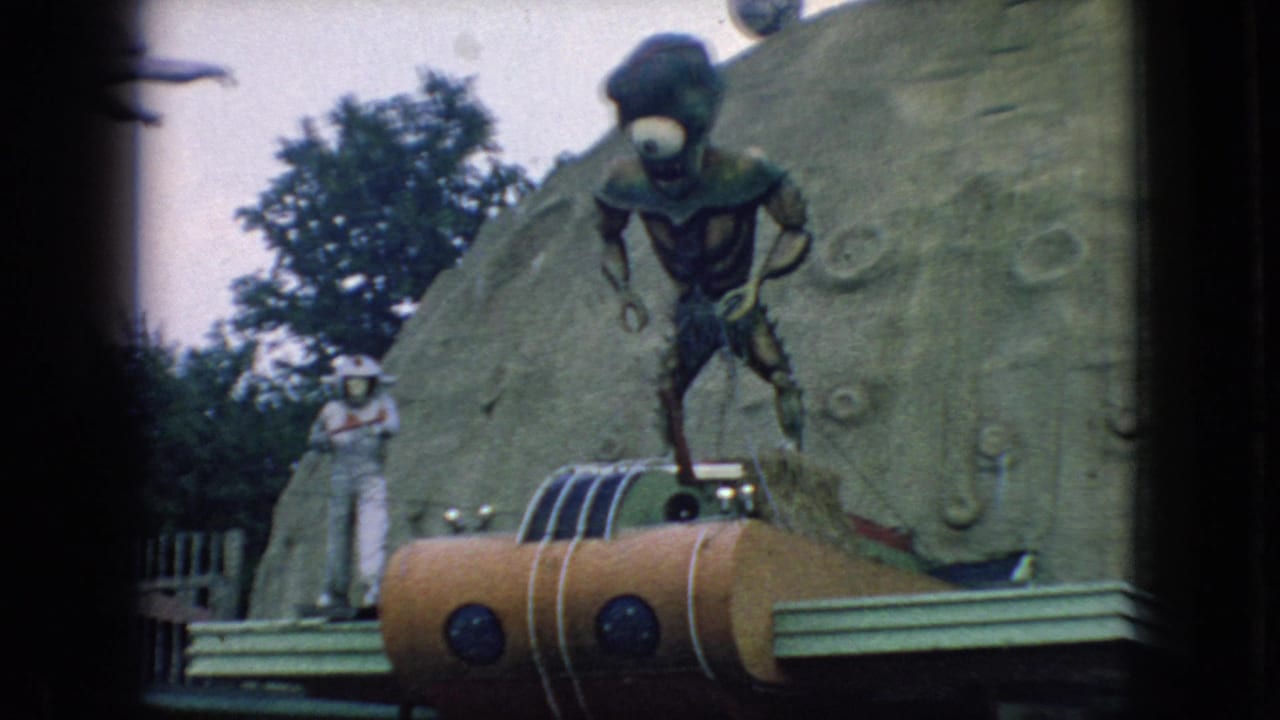 1966 New Jersey visitor-shot frame of a green one-eyed alien figure, a white astronaut figure, and an orange rocket set against a moon-like attraction backdrop.